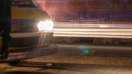 A blurred motion capture of a vehicle's headlights shining brightly on a road. Streaks of light trail across the image, indicating the movement of other vehicles in a nighttime setting. The background is dimly lit, creating a contrast with the bright lights.