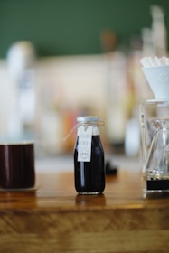 A small bottle labeled 'cold drip' sits on a wooden counter. To its left is a dark ceramic cup, and to the right is a glass pour-over coffee dripper with filters. The background is blurred, suggesting a coffee shop setting.