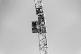 A black and white image depicting a large construction crane against a clear sky. The tower section of the crane, featuring beams and a control cabin, is visible.