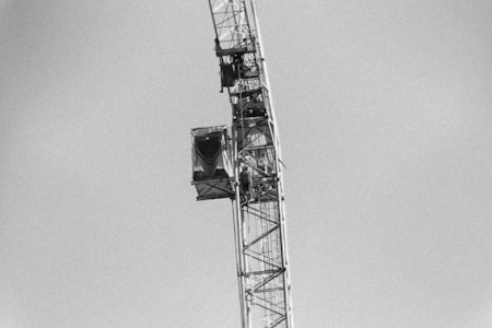 A black and white image depicting a large construction crane against a clear sky. The tower section of the crane, featuring beams and a control cabin, is visible.