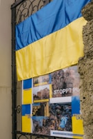 A Ukrainian flag is mounted on a black wrought iron structure next to a stone wall. Below the flag, there is a poster with several images, including scenes from Kharkiv and other places, possibly indicating conflict. Text on the poster includes phrases like 'STOPPT PUTIN' and 'PRAY FOR UKRAINE', suggesting a protest or solidarity message.