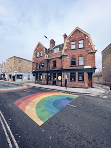 A street scene featuring a large old-style brick building with multiple gabled roofs and several windows. The building is located at a street corner. The road features a colorful rainbow painted on the asphalt, indicating an LGBTQ+ friendly area. A single person is seen walking along the sidewalk next to the building. The sky is overcast, adding a calm, subdued ambiance.