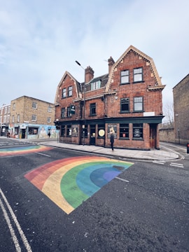 A street scene featuring a large old-style brick building with multiple gabled roofs and several windows. The building is located at a street corner. The road features a colorful rainbow painted on the asphalt, indicating an LGBTQ+ friendly area. A single person is seen walking along the sidewalk next to the building. The sky is overcast, adding a calm, subdued ambiance.