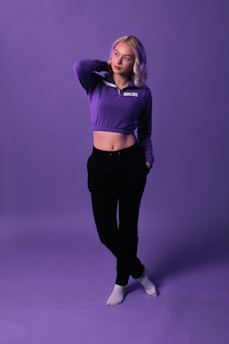 A person with light hair stands confidently against a purple backdrop. They are wearing a purple cropped hoodie with the word 'BOUJEE' on it, black pants, and white socks. The backdrop and attire create a cohesive aesthetic.