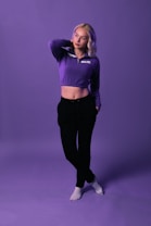 A person with light hair stands confidently against a purple backdrop. They are wearing a purple cropped hoodie with the word 'BOUJEE' on it, black pants, and white socks. The backdrop and attire create a cohesive aesthetic.