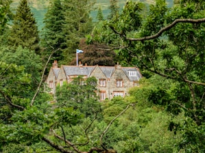 A stone house with a slate roof is nestled amidst a dense forest of lush green trees. The house has multiple chimneys and a Scottish flag flying on a pole. The architecture is traditional, and the house blends harmoniously with the natural surroundings.