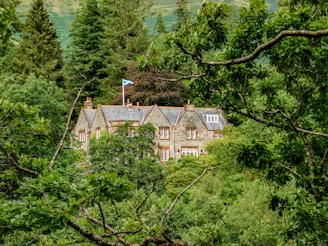 A stone house with a slate roof is nestled amidst a dense forest of lush green trees. The house has multiple chimneys and a Scottish flag flying on a pole. The architecture is traditional, and the house blends harmoniously with the natural surroundings.