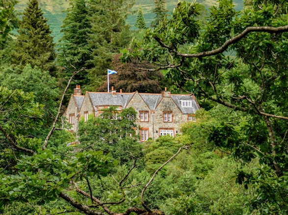 A stone house with a slate roof is nestled amidst a dense forest of lush green trees. The house has multiple chimneys and a Scottish flag flying on a pole. The architecture is traditional, and the house blends harmoniously with the natural surroundings.