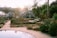 A serene garden setting with multiple greenhouses in the background, surrounded by various plants in raised beds. The sunlight casts a warm glow over the scene, and the garden paths are neatly paved with gravel. Trees and bushes add a sense of lushness, and a small pond is in the foreground reflecting the sky.