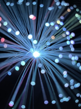 A burst of glowing fiber optic strands emitting light with varying shades, creating a vivid display of circular bokeh effects in different colors of blue, white, purple, and hints of red and green against a dark background.