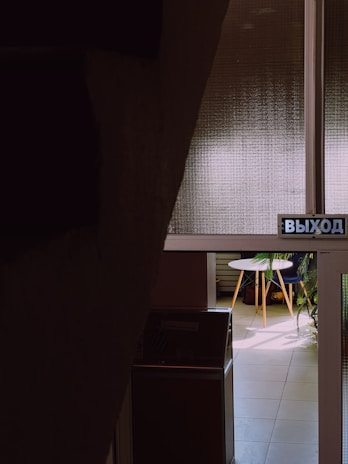 A dimly lit interior space featuring a partially visible room through a window. The room shows a small round table with light wood legs, surrounded by plants. A sign with Cyrillic text is present in the foreground.