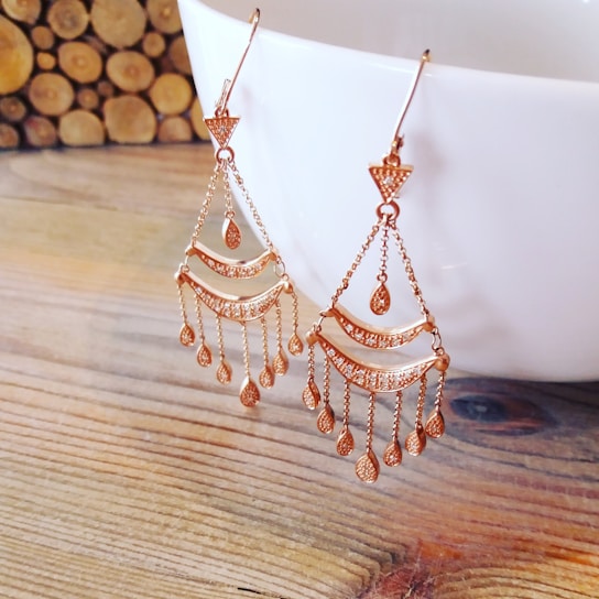 A pair of intricate gold chandelier earrings featuring multiple chain links with teardrop-shaped pendants. They are elegantly displayed hanging from a white ceramic cup placed on a wooden surface, providing a warm and rustic background.