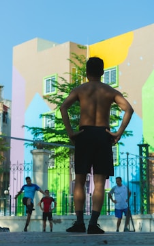 A group of people are engaged in a fitness activity outdoors. In the foreground, a man stands with his hands on his hips, facing away from the camera. In the background, other individuals are stretching or performing exercises. The setting is in front of a colorful, mural-covered building, with bright geometric patterns on the walls. A fence with decorative metalwork adds structure to the scene.
