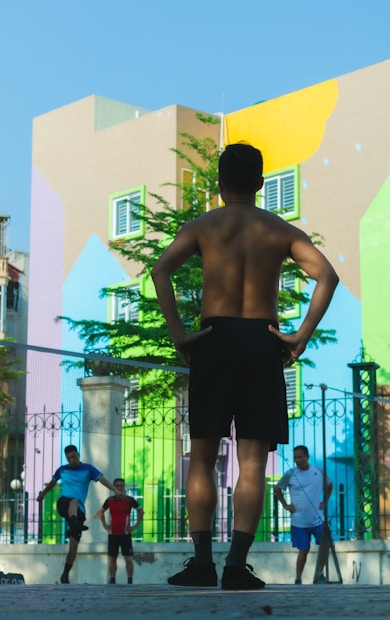 A group of people are engaged in a fitness activity outdoors. In the foreground, a man stands with his hands on his hips, facing away from the camera. In the background, other individuals are stretching or performing exercises. The setting is in front of a colorful, mural-covered building, with bright geometric patterns on the walls. A fence with decorative metalwork adds structure to the scene.