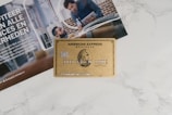 A gold American Express business credit card is placed on top of an advertisement featuring people in a professional setting. The surface is a white marble or similar texture. The card includes various numbers and text, with the notable American Express logo.