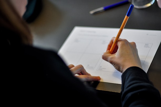 A person is seen writing on a paper placed on a dark surface. The hand is holding an orange pen, and the paper seems to contain organizational or planning layouts. Another pen, with a blue cap, is visible in the background.