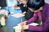 A woman in a purple shirt is writing on paper at a table, surrounded by others focused on similar tasks. Various colored pens and mugs are on the table, creating a professional and collaborative atmosphere.