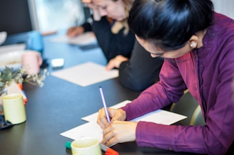 A woman in a purple shirt is writing on paper at a table, surrounded by others focused on similar tasks. Various colored pens and mugs are on the table, creating a professional and collaborative atmosphere.