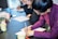 A woman in a purple shirt is writing on paper at a table, surrounded by others focused on similar tasks. Various colored pens and mugs are on the table, creating a professional and collaborative atmosphere.