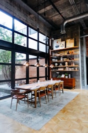 A stylish dining area with a wooden table and eight matching chairs set on a large rug. The space features large windows allowing natural light to flood in, with views of a brick wall and courtyard outside. Hanging lights and industrial piping add a modern touch. A bookshelf and cabinet stand on the right, displaying art, vases, and decorative items.