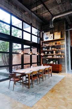 A stylish dining area with a wooden table and eight matching chairs set on a large rug. The space features large windows allowing natural light to flood in, with views of a brick wall and courtyard outside. Hanging lights and industrial piping add a modern touch. A bookshelf and cabinet stand on the right, displaying art, vases, and decorative items.