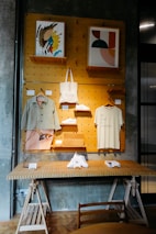 A stylish display shelf against a dark textured wall features two pieces of abstract artwork, a canvas tote bag, a shirt with 'Be kind to strangers.' printed on it, and a pair of white sneakers. The setup includes wooden shelves and hangers, with additional items like a lamp and small boxes on a table below.