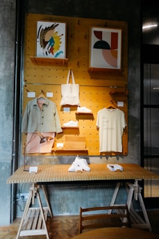 A stylish display shelf against a dark textured wall features two pieces of abstract artwork, a canvas tote bag, a shirt with 'Be kind to strangers.' printed on it, and a pair of white sneakers. The setup includes wooden shelves and hangers, with additional items like a lamp and small boxes on a table below.