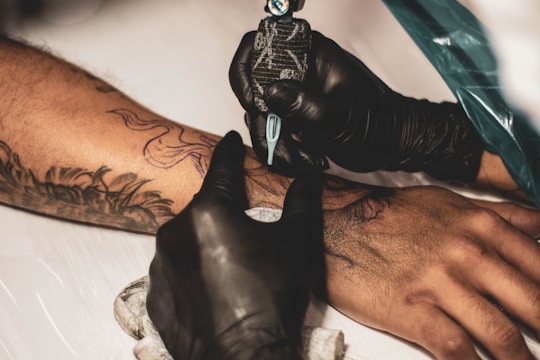 A tattoo artist wearing black gloves is applying ink on a person's forearm. The forearm already has an outline sketch and appears to be in the process of being filled in. The artist uses a tattoo machine, and the scene is set on a white surface.