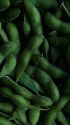 A close-up view of green edamame pods piled together. The texture appears fuzzy, and the pods are slightly curved, showcasing their natural form.