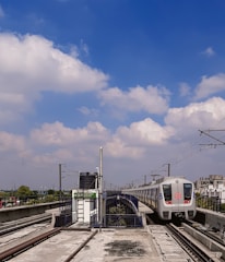 A modern metro train is approaching a station with electrified tracks on an elevated platform. The train is white with blue and red markings, set against a backdrop of a clear blue sky with scattered white clouds. Various infrastructure elements, such as electric poles and a sign with station information, are visible.