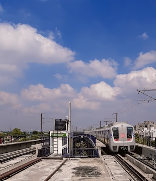 A modern metro train is approaching a station with electrified tracks on an elevated platform. The train is white with blue and red markings, set against a backdrop of a clear blue sky with scattered white clouds. Various infrastructure elements, such as electric poles and a sign with station information, are visible.