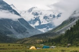 A serene valley surrounded by tall, snow-covered mountains partially shrouded in clouds with a grassy meadow in the foreground featuring several colorful tents. Lush greenery lines the valley, adding to the natural tranquility of the scene.
