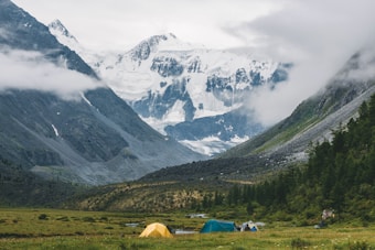 A serene valley surrounded by tall, snow-covered mountains partially shrouded in clouds with a grassy meadow in the foreground featuring several colorful tents. Lush greenery lines the valley, adding to the natural tranquility of the scene.