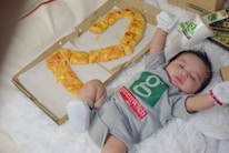 A baby in a gray outfit lies on a fluffy white surface, surrounded by pizza boxes. The baby is wearing a Greenwich-branded onesie and is stretching arms outward. A box is partially open with pizza slices arranged in the shape of a number, possibly representing an age or occasion. A cup with the Greenwich logo is placed nearby.