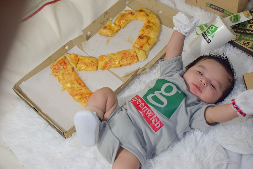 A baby in a gray outfit lies on a fluffy white surface, surrounded by pizza boxes. The baby is wearing a Greenwich-branded onesie and is stretching arms outward. A box is partially open with pizza slices arranged in the shape of a number, possibly representing an age or occasion. A cup with the Greenwich logo is placed nearby.