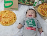 A baby lies on a soft, white furry surface, dressed in a gray onesie with a green logo and the word 'greenwich.' The baby is smiling with closed eyes. There are two pizzas on either side, one topped with cheese and another with ham and pineapple, placed near paper bags and boxes with similar branding.