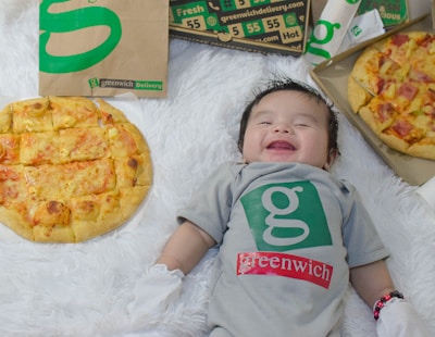 A baby lies on a soft, white furry surface, dressed in a gray onesie with a green logo and the word 'greenwich.' The baby is smiling with closed eyes. There are two pizzas on either side, one topped with cheese and another with ham and pineapple, placed near paper bags and boxes with similar branding.