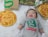 A baby lies on a soft, white furry surface, dressed in a gray onesie with a green logo and the word 'greenwich.' The baby is smiling with closed eyes. There are two pizzas on either side, one topped with cheese and another with ham and pineapple, placed near paper bags and boxes with similar branding.