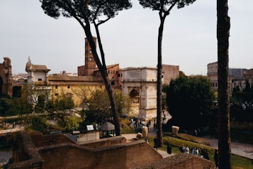 An ancient historical site with large stone structures and arches, surrounded by lush greenery and tall trees. A mix of visitors and tourists are scattered around, exploring the area. The architecture is reminiscent of classic Roman style, with one structure resembling the Colosseum visible in the background.