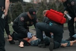 Several emergency responders attend to a person lying on the ground. One responder kneels beside the individual, holding medical equipment, while another is positioned nearby with a red medical bag. The scene suggests a medical emergency, with responders wearing uniforms and equipment indicating a professional, urgent atmosphere.