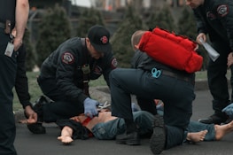 Several emergency responders attend to a person lying on the ground. One responder kneels beside the individual, holding medical equipment, while another is positioned nearby with a red medical bag. The scene suggests a medical emergency, with responders wearing uniforms and equipment indicating a professional, urgent atmosphere.