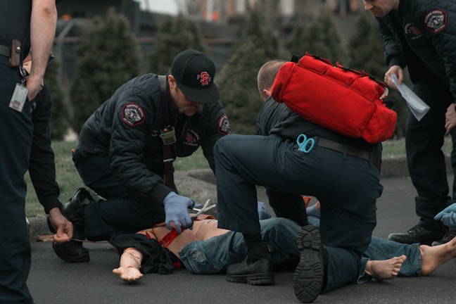 Several emergency responders attend to a person lying on the ground. One responder kneels beside the individual, holding medical equipment, while another is positioned nearby with a red medical bag. The scene suggests a medical emergency, with responders wearing uniforms and equipment indicating a professional, urgent atmosphere.
