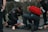 Several emergency responders attend to a person lying on the ground. One responder kneels beside the individual, holding medical equipment, while another is positioned nearby with a red medical bag. The scene suggests a medical emergency, with responders wearing uniforms and equipment indicating a professional, urgent atmosphere.