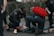 Several emergency responders attend to a person lying on the ground. One responder kneels beside the individual, holding medical equipment, while another is positioned nearby with a red medical bag. The scene suggests a medical emergency, with responders wearing uniforms and equipment indicating a professional, urgent atmosphere.