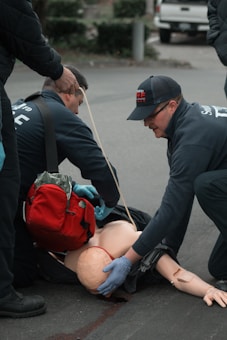Emergency responders in uniforms are performing a training exercise with a medical mannequin on the pavement. There is a red medical bag near the mannequin, and one responder is wearing blue gloves.
