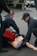Emergency responders in uniforms are performing a training exercise with a medical mannequin on the pavement. There is a red medical bag near the mannequin, and one responder is wearing blue gloves.