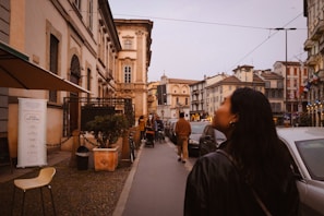 A city street lined with historic buildings, with a person walking in the foreground. The street is bustling with people and cars parked along the sides. The architecture features ornate details and vibrant colors. A café or restaurant appears on the left, with outdoor seating and a menu displayed.
