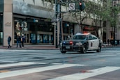 A police car with lights flashing is parked on a city street. Several pedestrians are standing on a sidewalk near an intersection, next to a building with multiple AT&T banners. Trees line the street and a one-way sign is visible.
