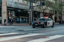 A police car with lights flashing is parked on a city street. Several pedestrians are standing on a sidewalk near an intersection, next to a building with multiple AT&T banners. Trees line the street and a one-way sign is visible.