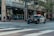 A police car with lights flashing is parked on a city street. Several pedestrians are standing on a sidewalk near an intersection, next to a building with multiple AT&T banners. Trees line the street and a one-way sign is visible.